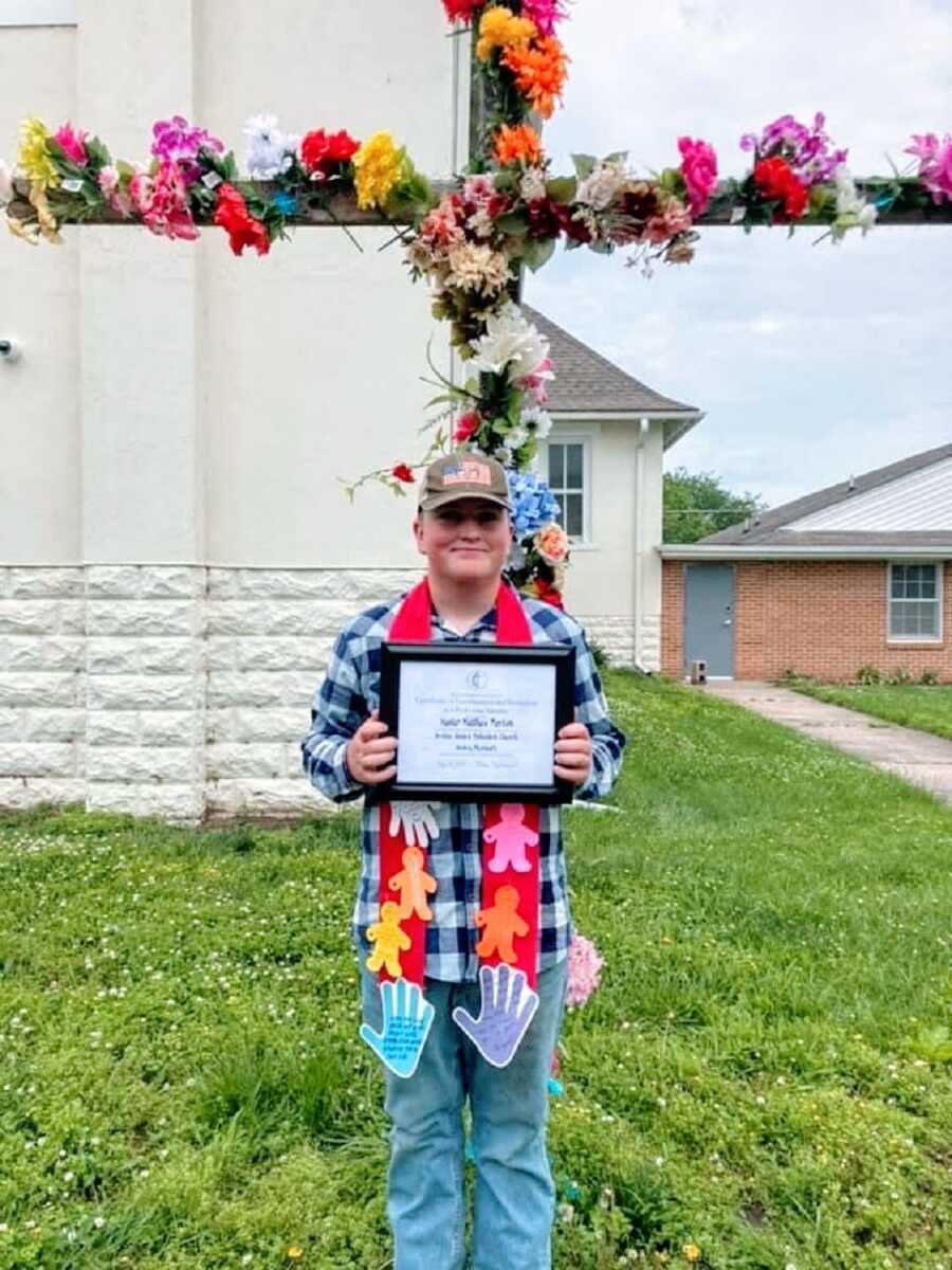 A boy stands in front of a cross covered in flowers