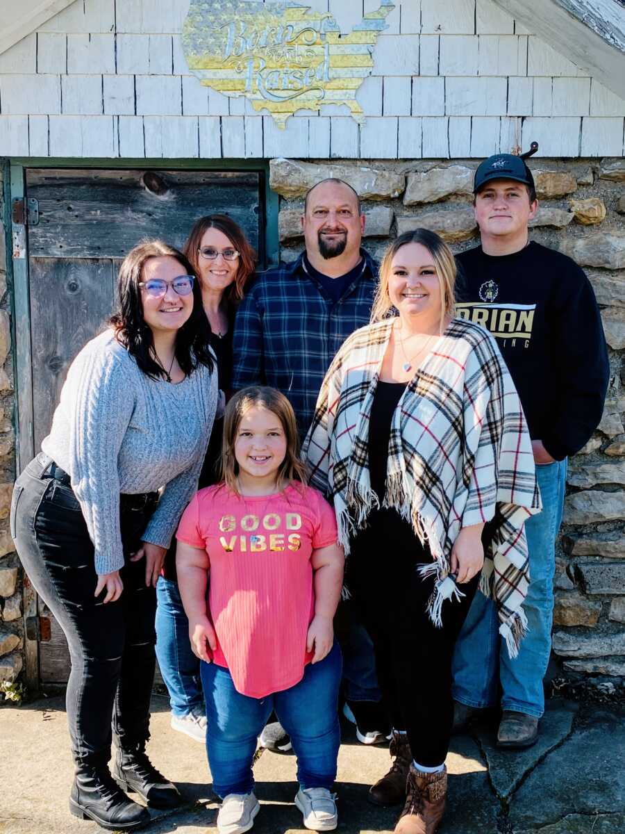 A family of six stand together against a wall