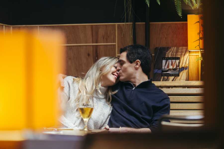 Wife leans into husband's shoulder and he gently kisses her nose while they sit together in a cozy room with a glass of wine on the table in front of them.