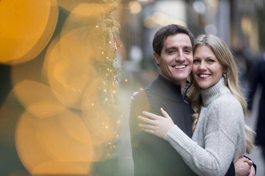 Husband and wife embrace one another in winter picture with blurry lights out of focus.