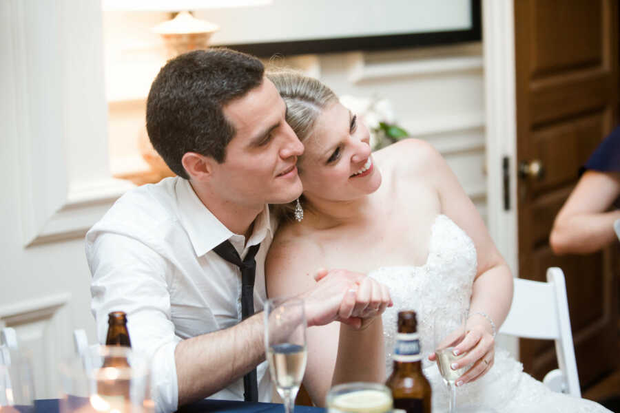 Husband and wife sit together on their wedding day holding one another's hands as wife leans into husband.