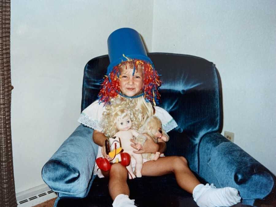 Young girl sits in armchair holding baby dolls with a blue bucket on top of her head.