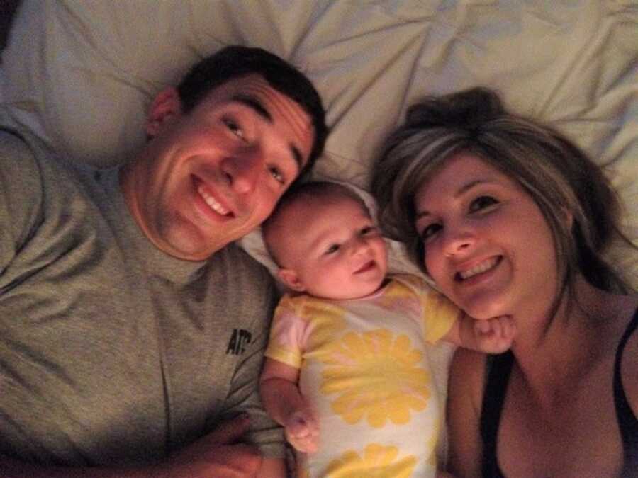Young couple lie on bed with baby girl between them, smiling up at the camera.