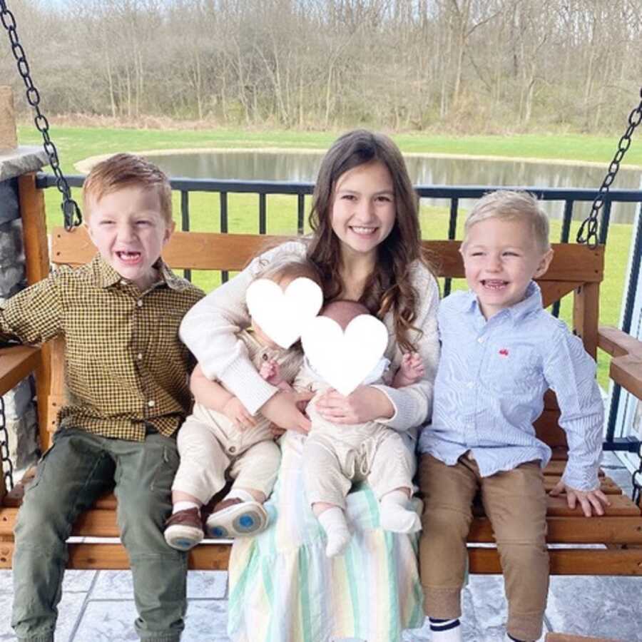 Adoptive siblings sit on porch swing holding foster babies.