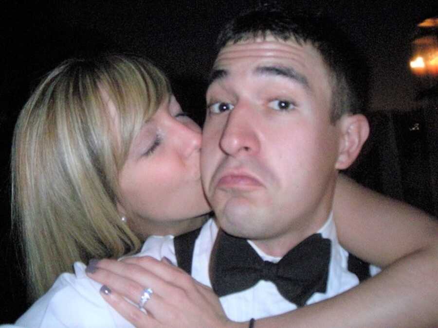 Woman wraps her arm around man in a tuxedo and kisses him on the cheek, showing off engagement ring on the night of his proposal.