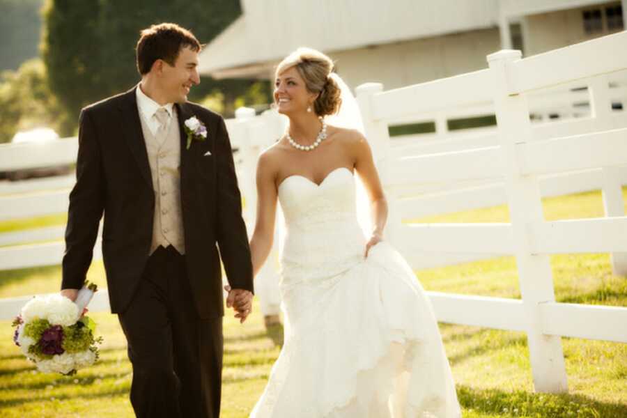 Young couple holding hands and wearing wedding clothes take picture outside in front of white fence.