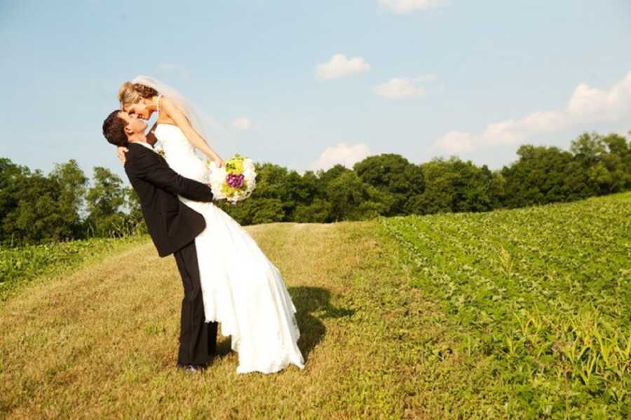 Groom lifts up bride and kisses her in an open field.