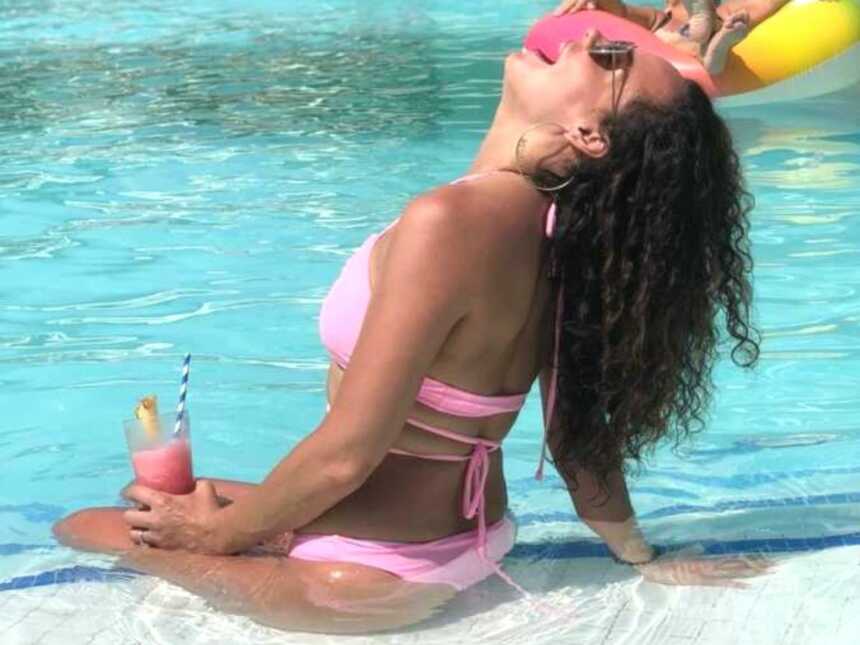 A woman sits by the pool holding a drink and wearing a bikini