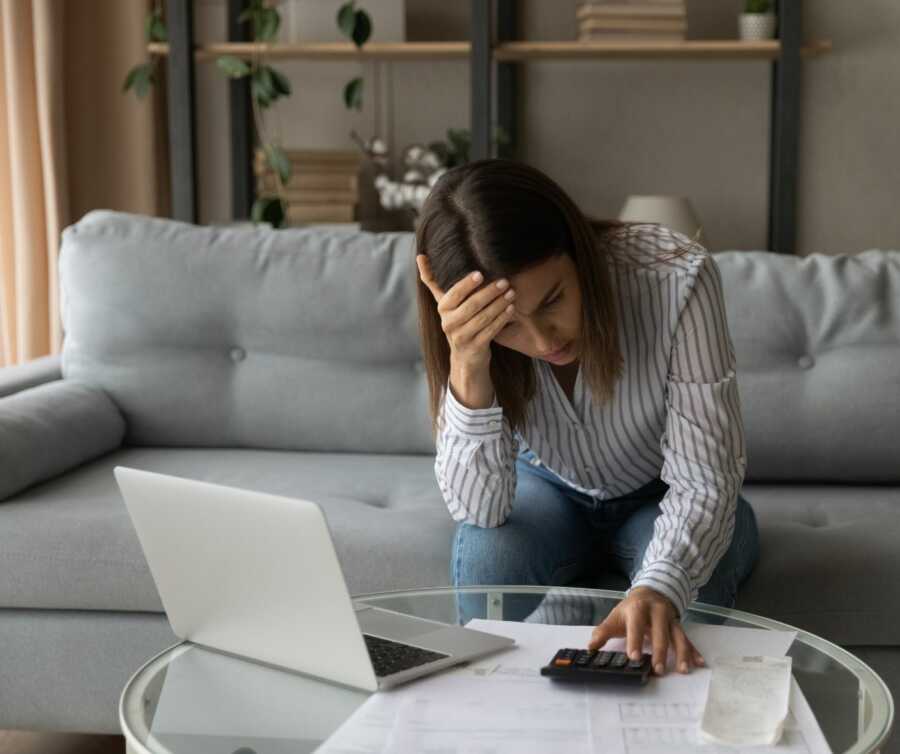 Woman sits on couch with her head in her hand as she pours over bills and calculates costs.