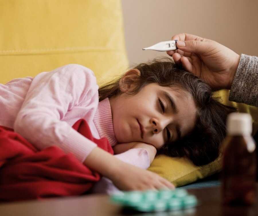 Little girl sleeps on yellow pillow while a woman's hand can be seen holding a thermometer.