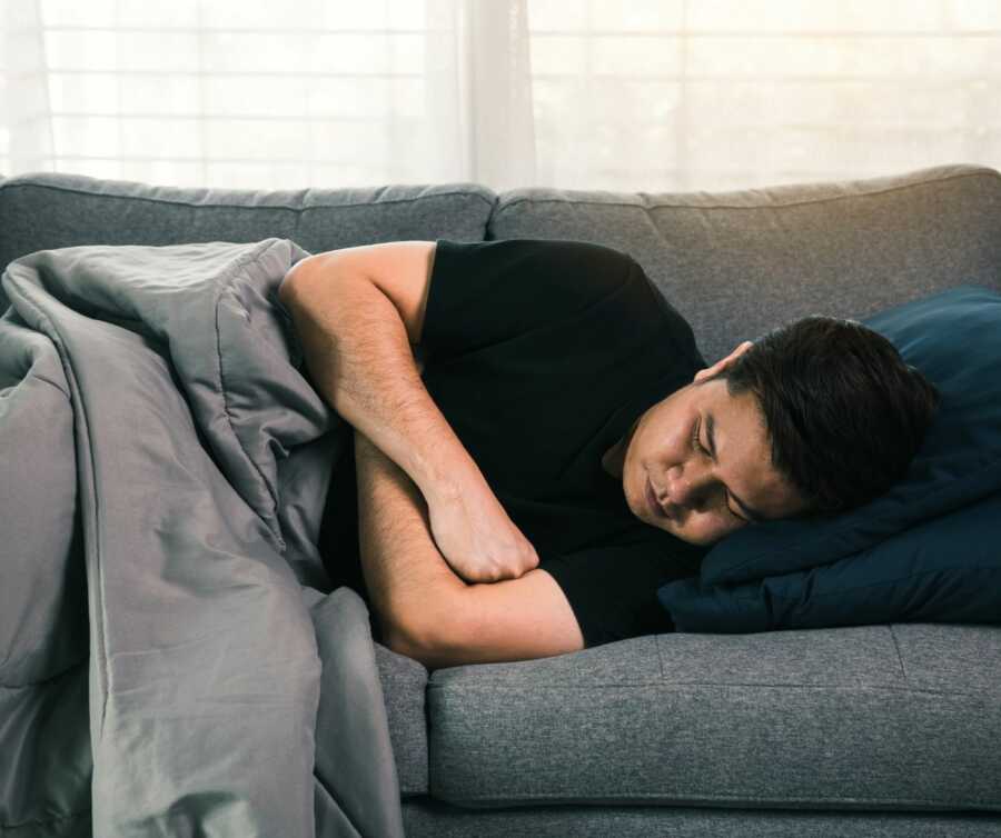 Asian man sleeps on couch with arms folded against chest and grey blanket keeping him warm.