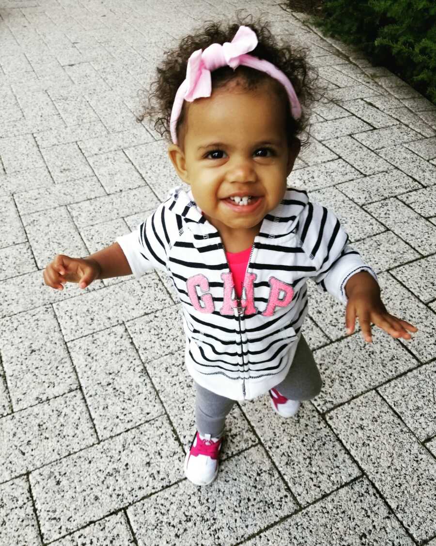 Black toddler girl in foster care smiles for camera, wearing a striped jacket and pink shirt.