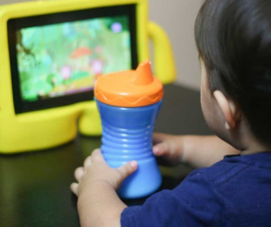 Young toddler holds sippy cup while watching cartoons on a children's screen device.