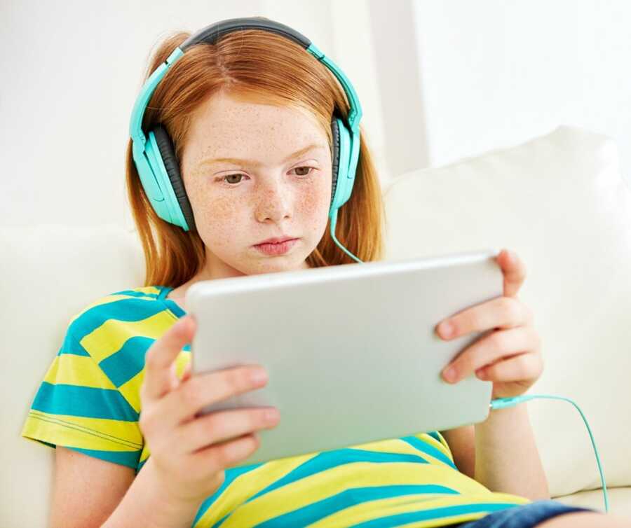 Red headed girl wearing yellow and blue striped shirt sits on the couch wearing headphones and watching her device.