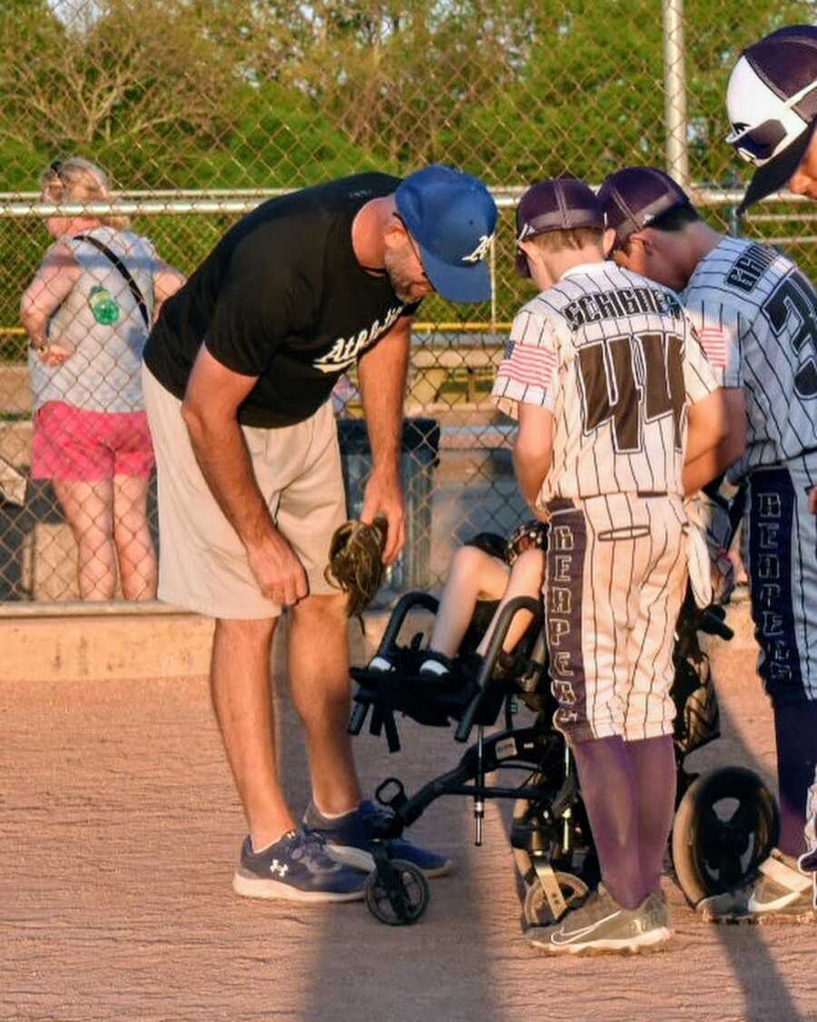 baseball coach and players bend over boy in wheelchair to talk to him
