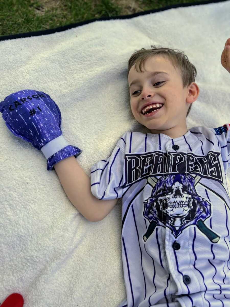 young boy with disability lays on ground in baseball jersey smiling wide
