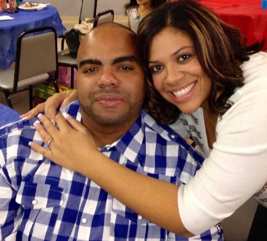 Military wife puts arms around husbands shoulders and smiles with her face next to his.