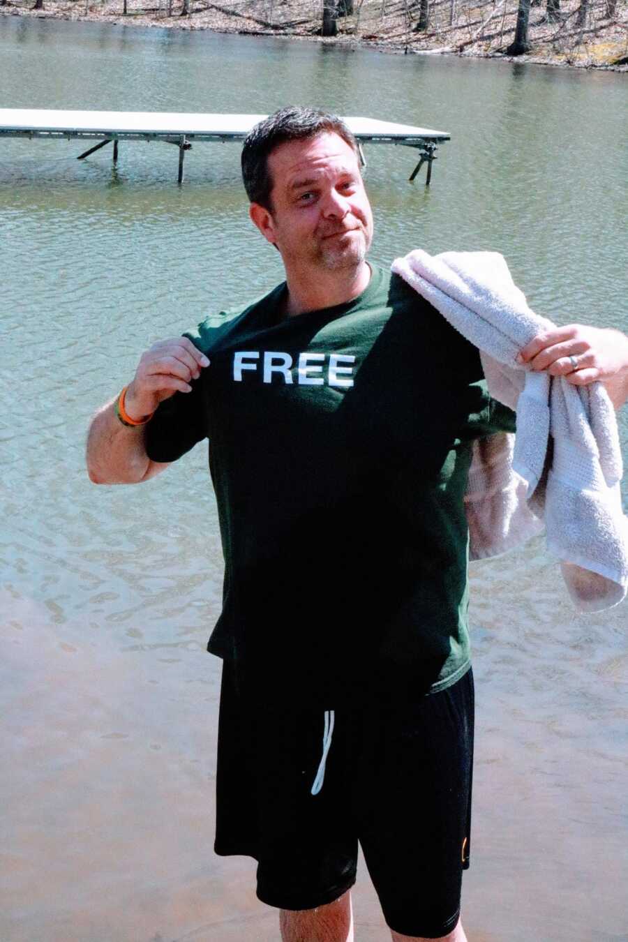 A man wearing a green T-shirt reading FREE holding a towel by the water