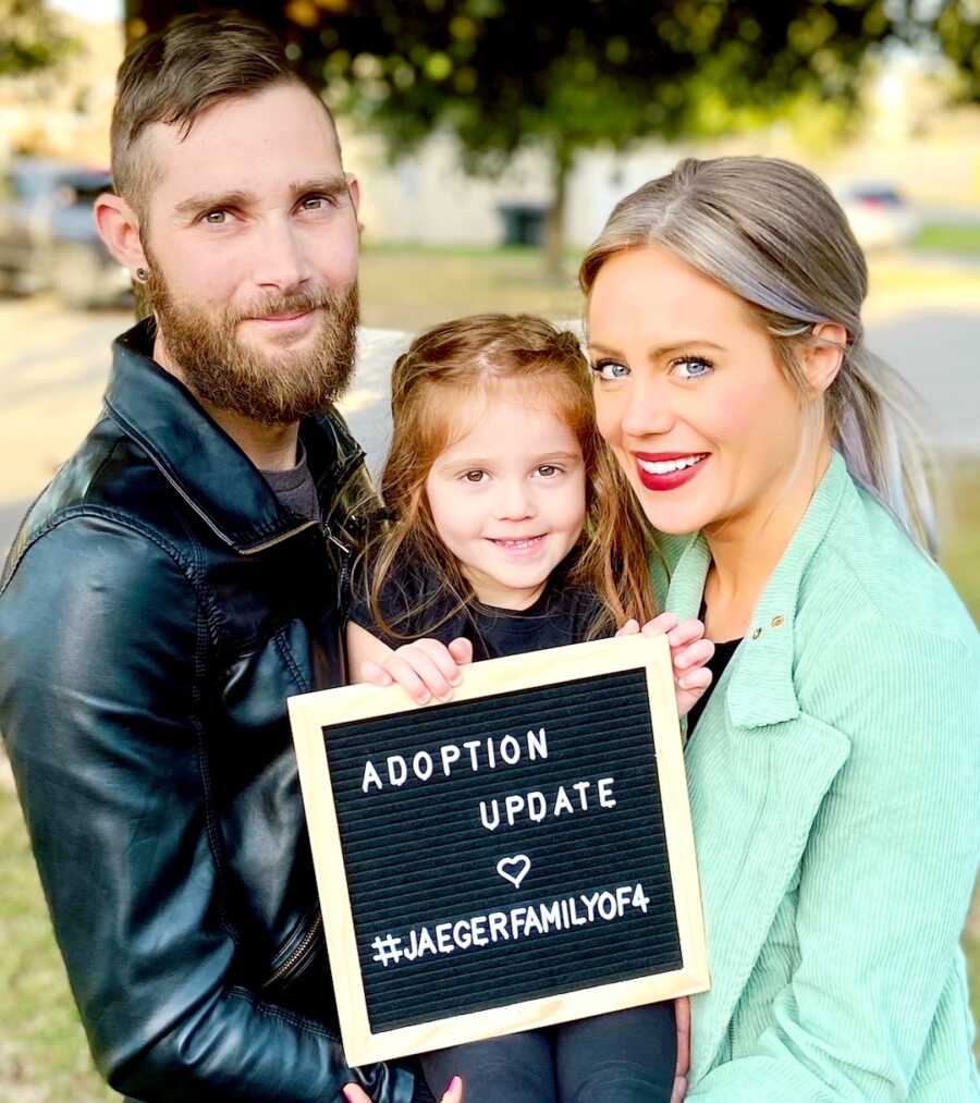 family of three stand holding letter sign sharing adoption announcement