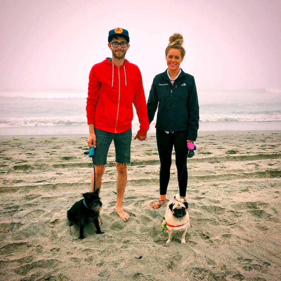couple stands on beach holding hands holding their two dogs