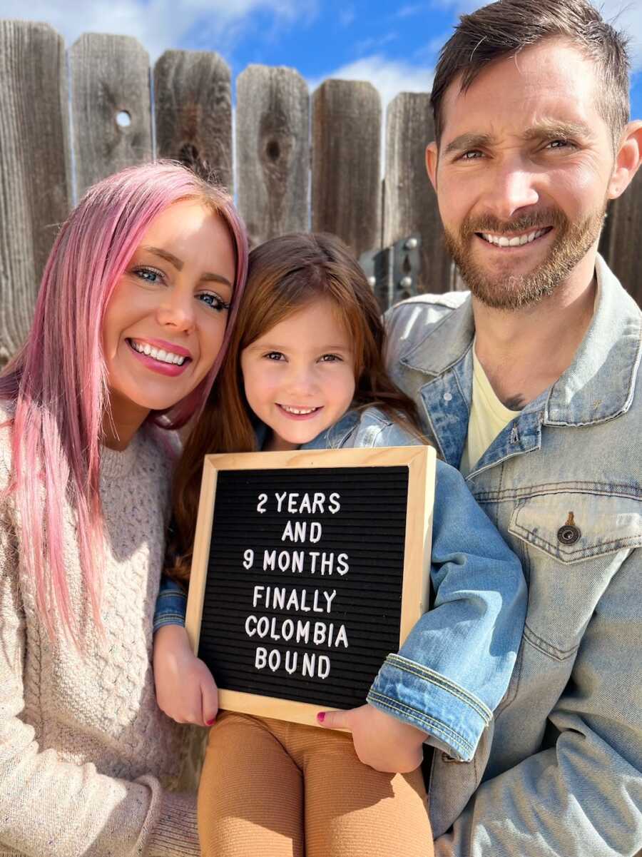 family of three stands holding letter sign sharing they are leaving for Colombia for adoption