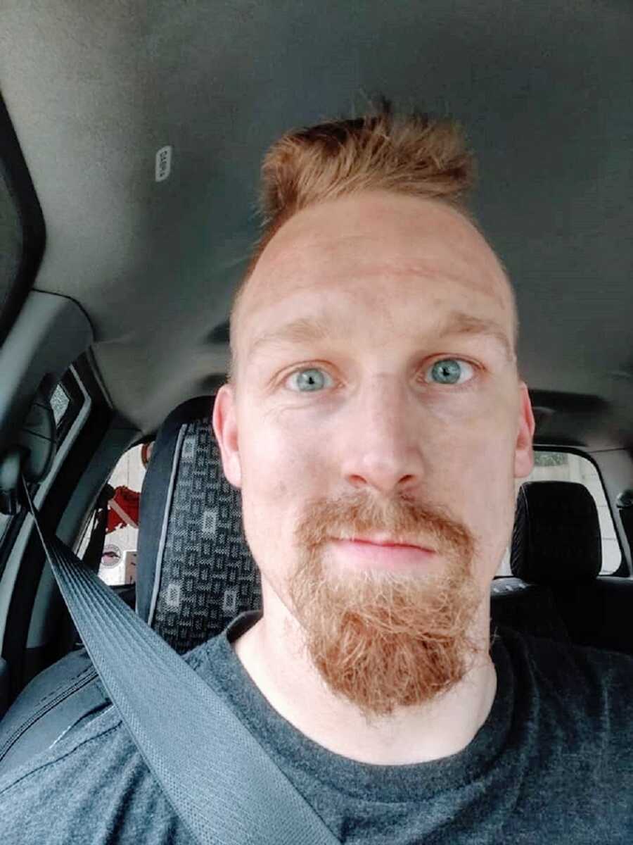 A man with a goatee and grey eyes sits in his car