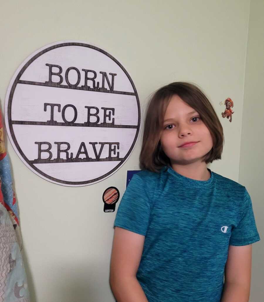 boy in blue shirt sitting next to a sign that says born to be brave