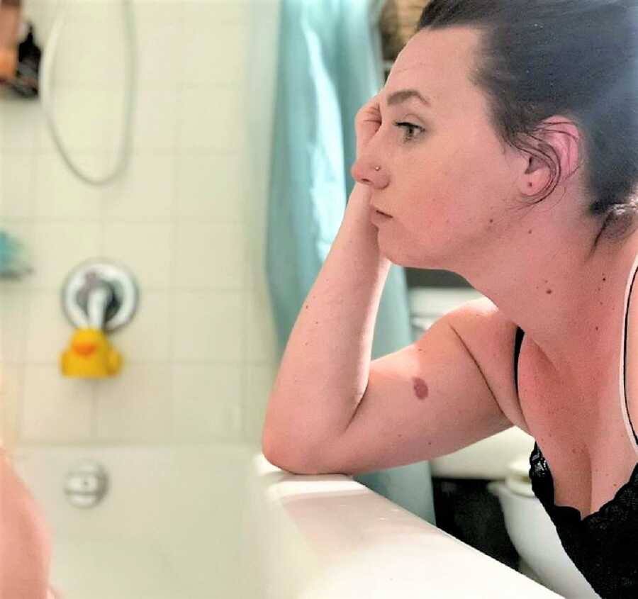 exhausted-looking mom resting her arm on the bathtub while observing her child take a bath