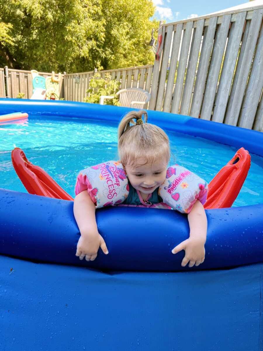 Little girl swims in an above-ground pool in her backyard with pink floaties on