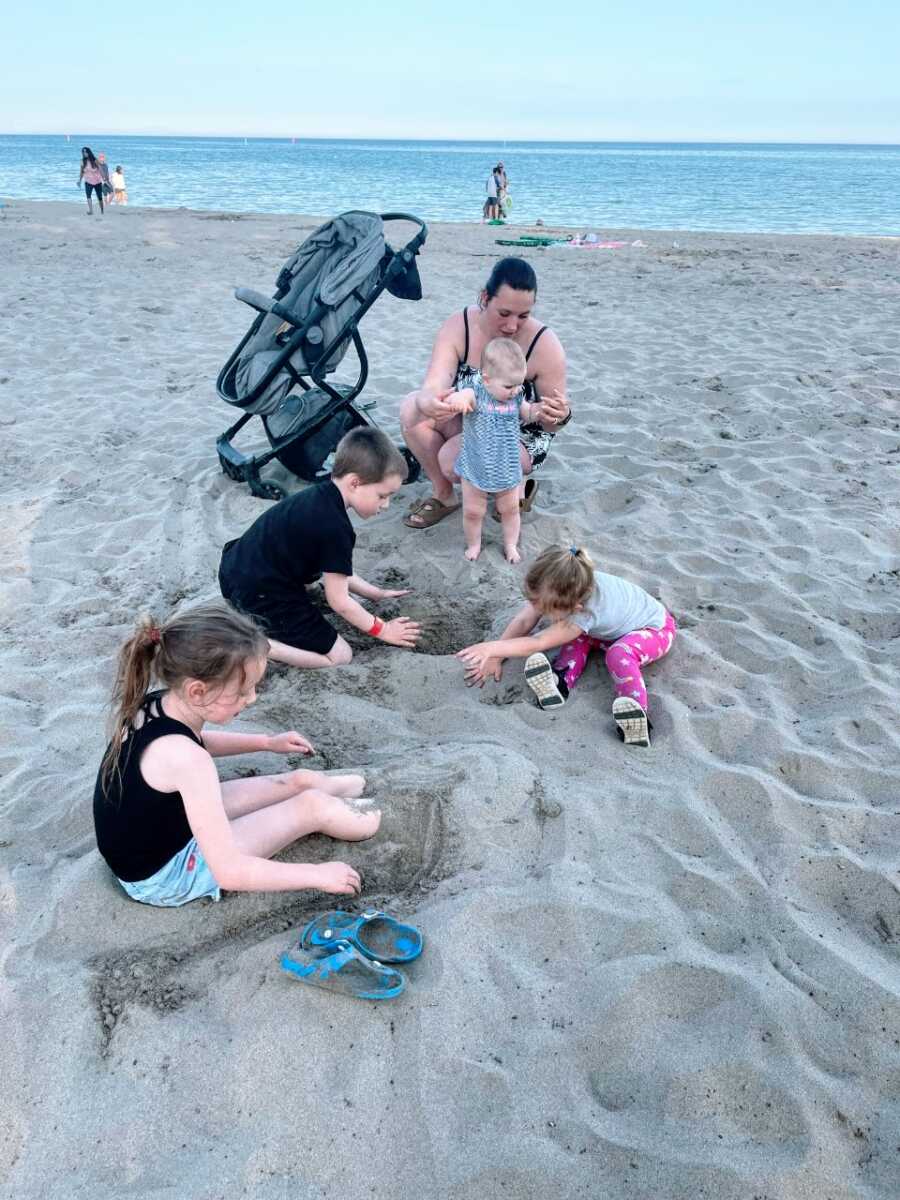 Mom sits down at the beach with her four kids while they all build a sandcastle together