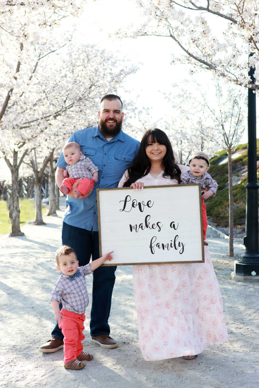 portrait of husband and wife holding their 3 adoptive sons while holding a sing that says "LOVE MAKES A FAMILY"