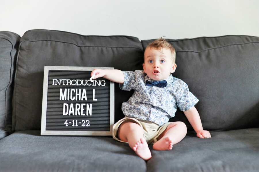 Baby boy wearing a bowtie and sitting on a couch holding a sign with his name 