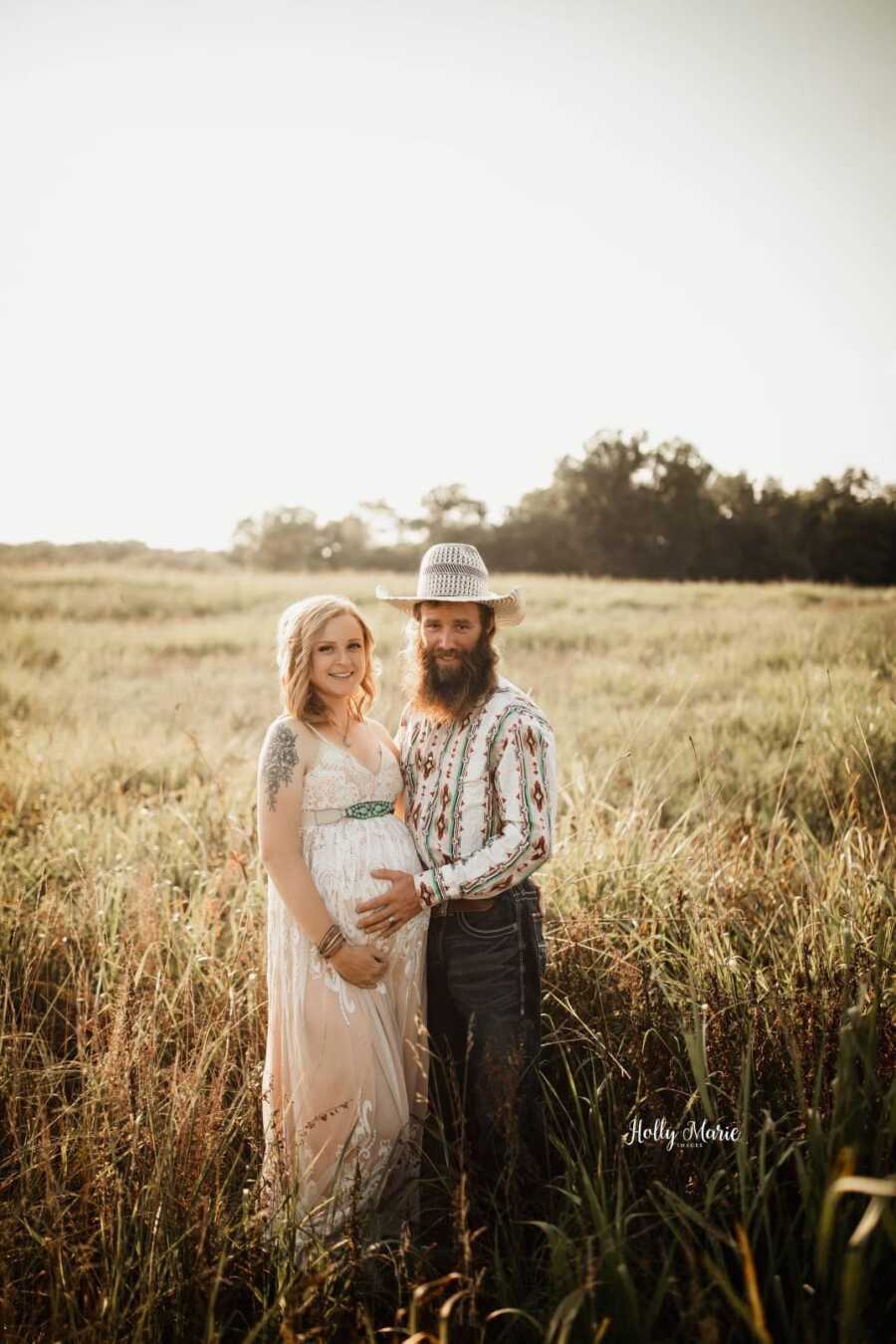 pregnant wife wearing a white long dress next to her husband who is holding her baby bump 