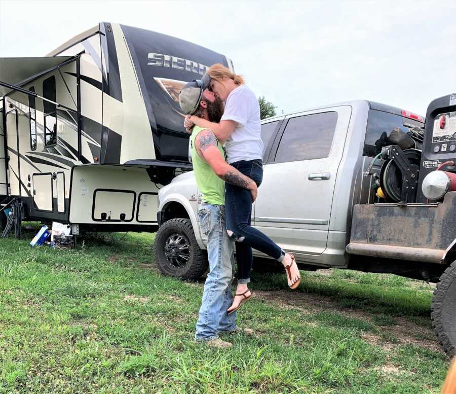 Husband carrying his wife on his arm while giving her a kiss in front of their camper house