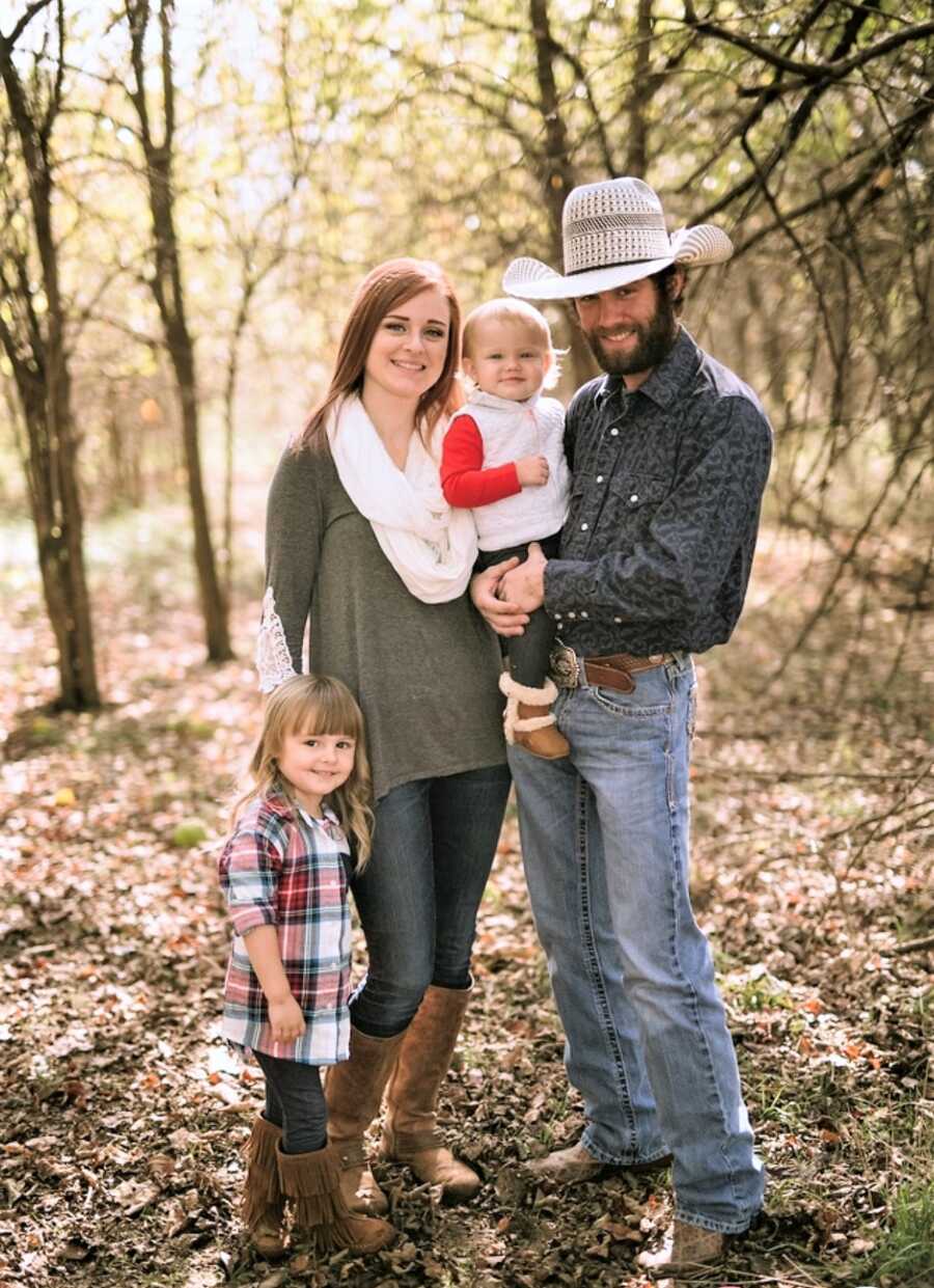 husband and wife standing in the woods next to their little daughter while holding their baby daughter 
