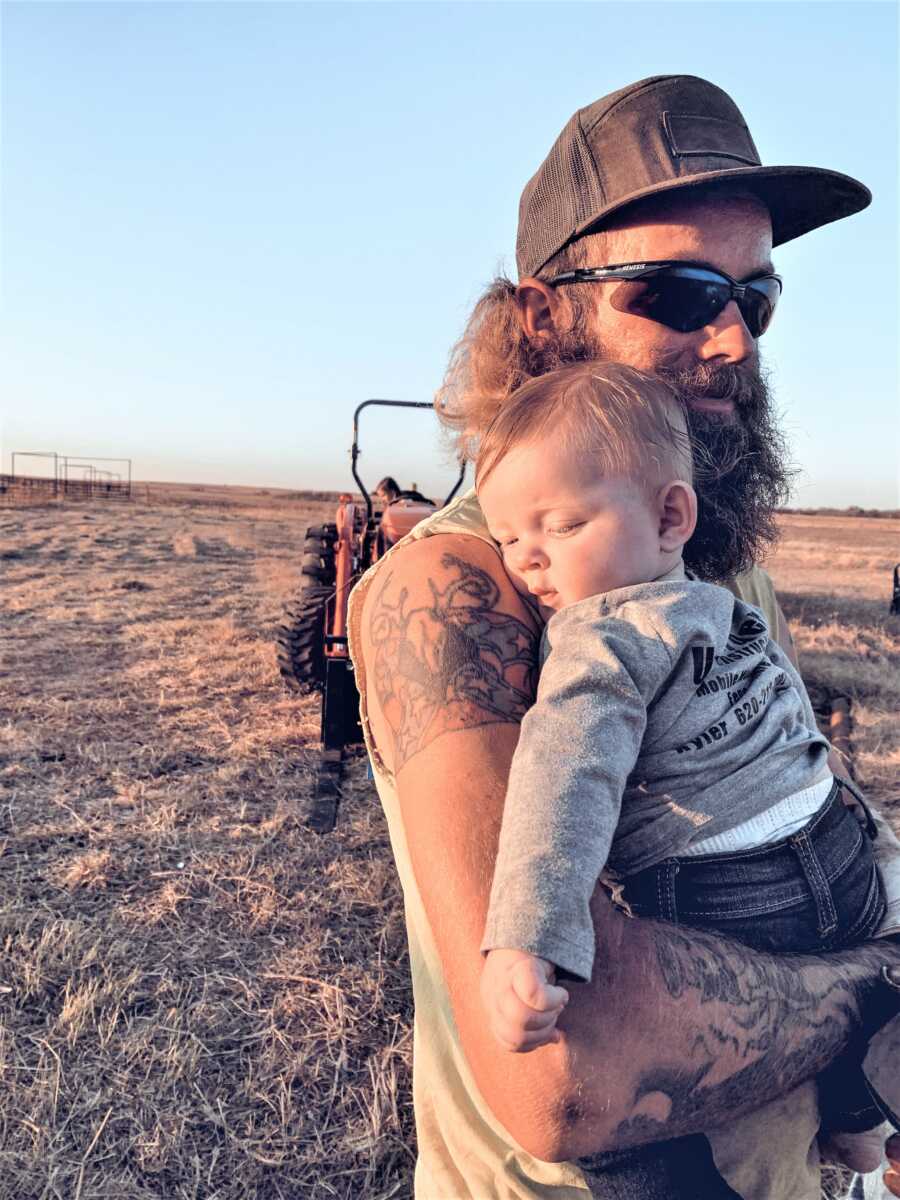 dad holding his baby son at a farm with a tractor in the back 