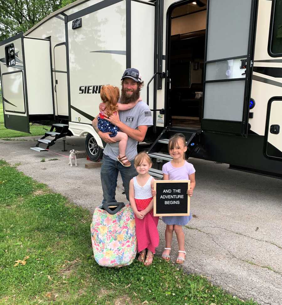 dad and three daughters standing in front of their camper house holding a sign that says "AND THE ADVENTURE BEGINS"