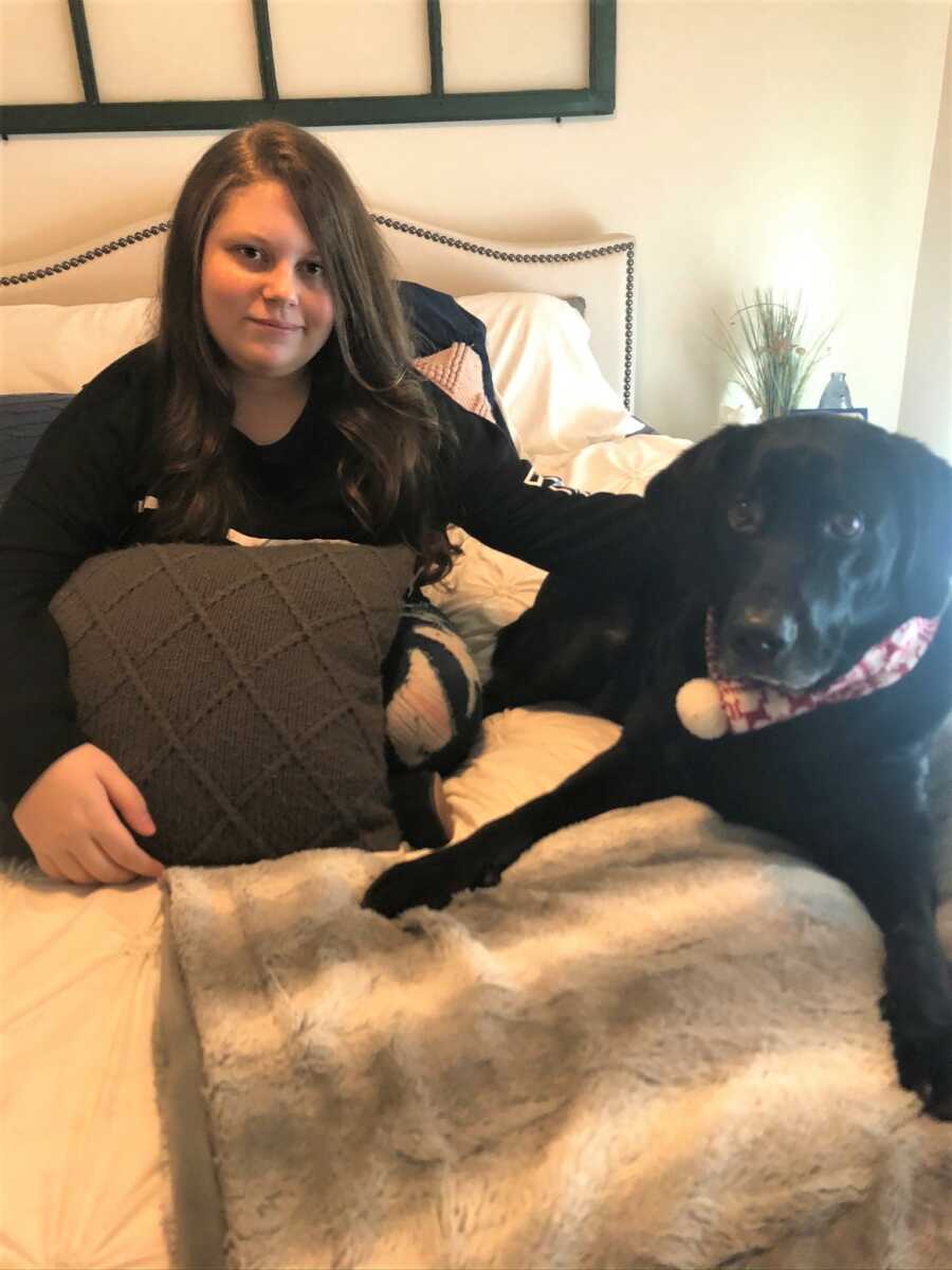 Teen foster girl sits on bed with hand resting on family black lab.