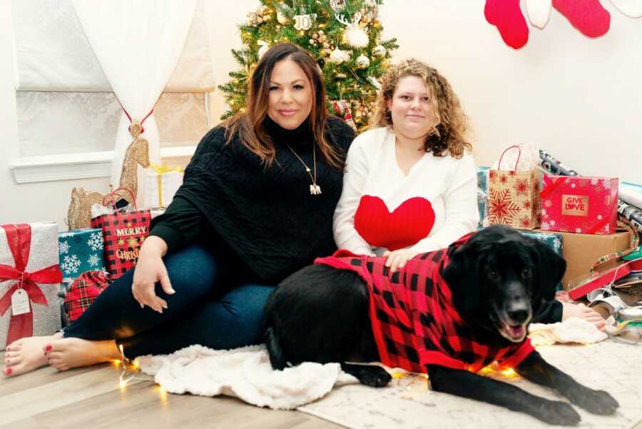 Single adoptive mom and teen daughter take picture with family dog in front of Christmas tree and presents.