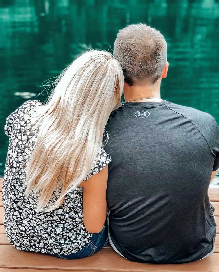 Husband and wife sitting in front of a lake while wife rests her head on her husband's shoulder