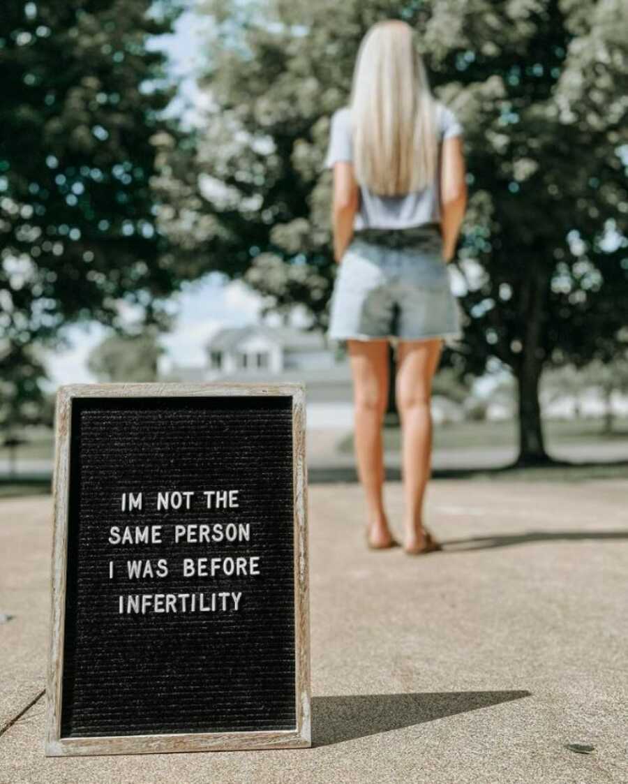 Woman, unfocused, stands in the distance with a sign about her infertility journey focused in the center of the image