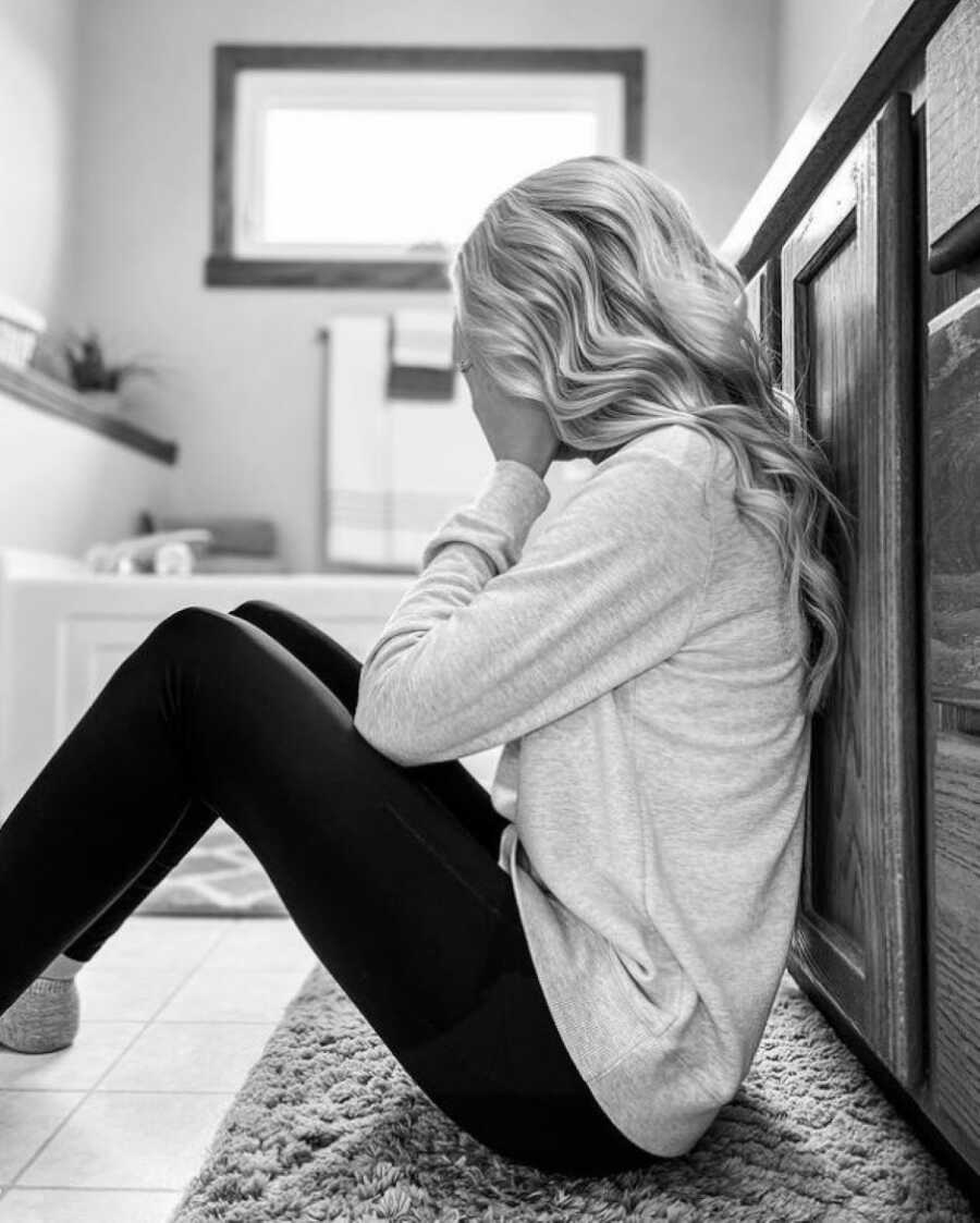 Woman battling infertility sits on the floor of her bathroom with her head in her hands