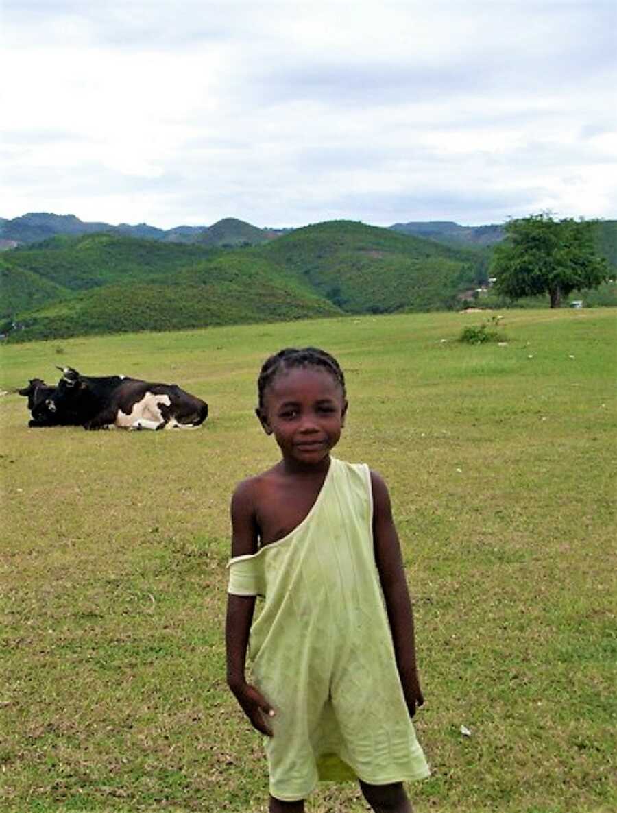 Young Haitian girl standing in a grass field wearing a green jumper with mountains and cows in the background 