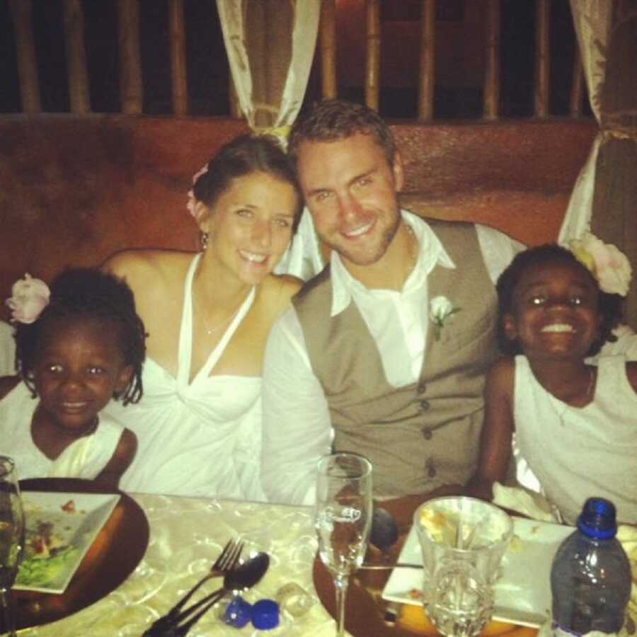 American man and woman on their wedding celebration with their two adoptive, Haitian daughters sitting next to them 
