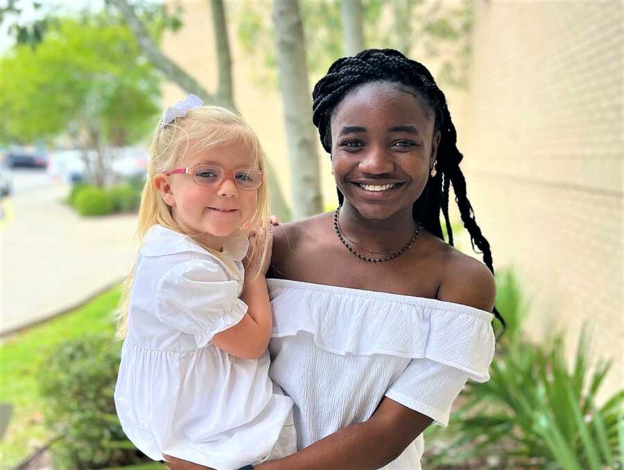 Older sister holding her younger sister in her arms while both wear white outfits 