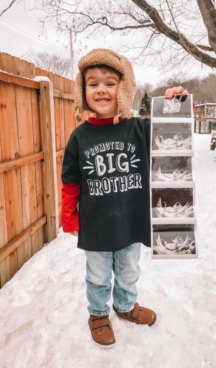 Little boy announces his mom's pregnancy by holding ultrasound photos while wearing a "promoted to big brother" T-shirt