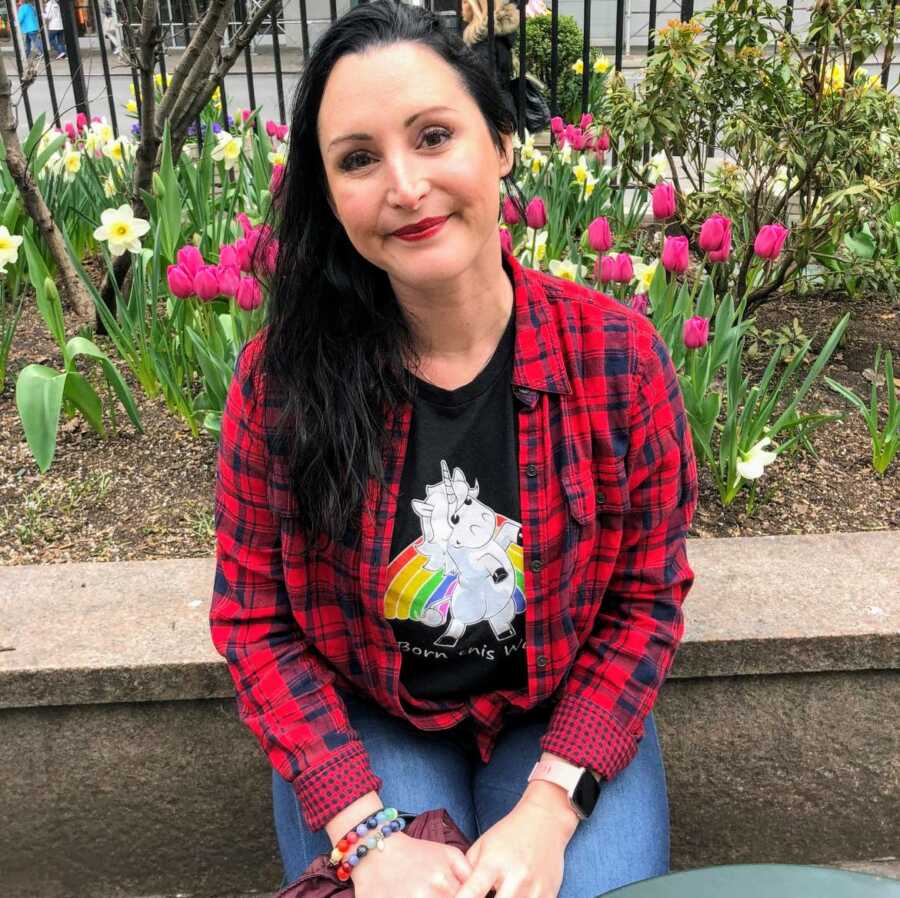 Bisexual person sits on a concrete ledge in a rainbow t-shirt with a unicorn on it that reads "born this way"