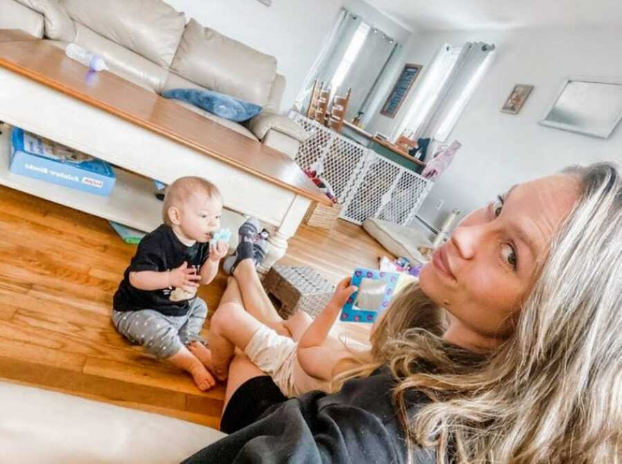 Girl mom takes a selfie on the couch while one daughter sits in her lap and the other sits on the coffee table