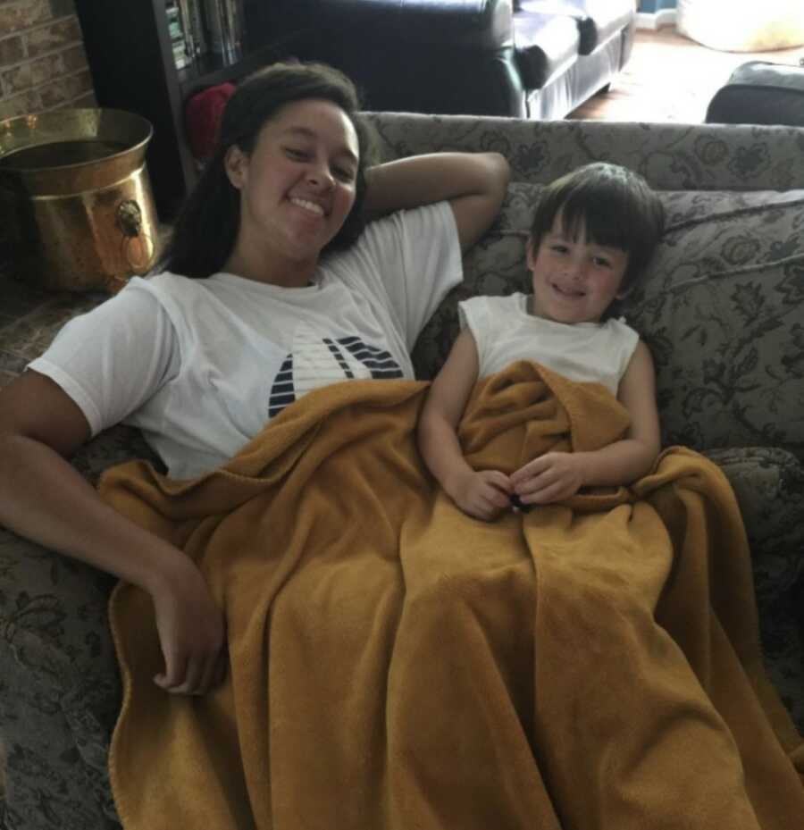 Young woman in foster care loves having a baby brother and sits on the couch sharing a blanket with him.