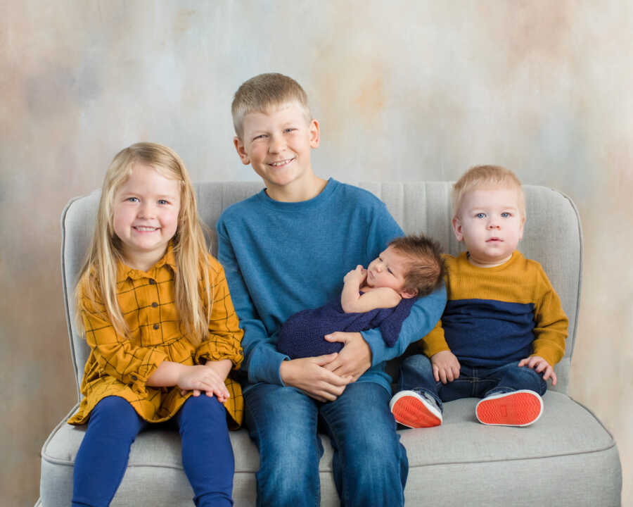 Siblings sit on couch with oldest holding brand new baby brother.